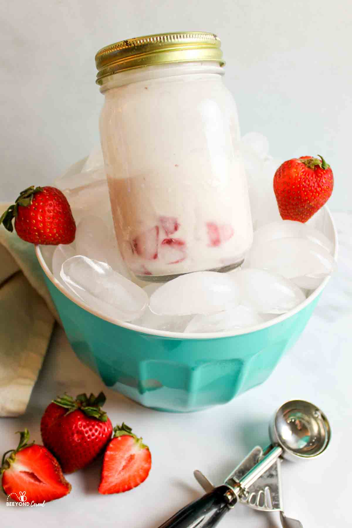 a jar of strawberry ice cream in a bowl of ice next to frehs strawberries and an ice cream scoop