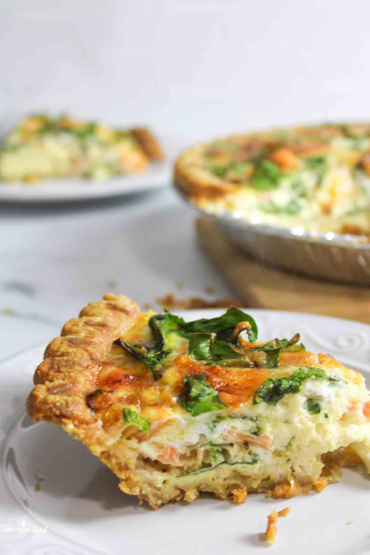 a slice of Salmon and Spinach Quiche on a white plate with more in the background