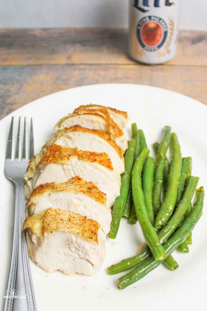 a fork, chicken breast and green beans on a plate with a can of beer in the background
