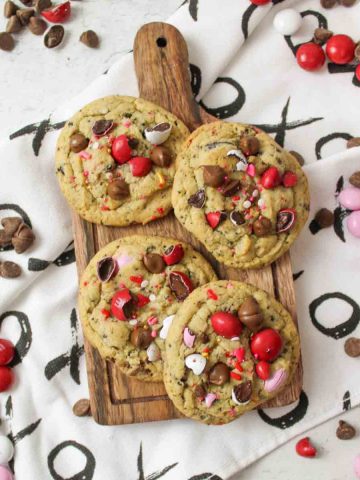 4 cookies on a wooden board with scattered M&Ms and chocolate chips around it.