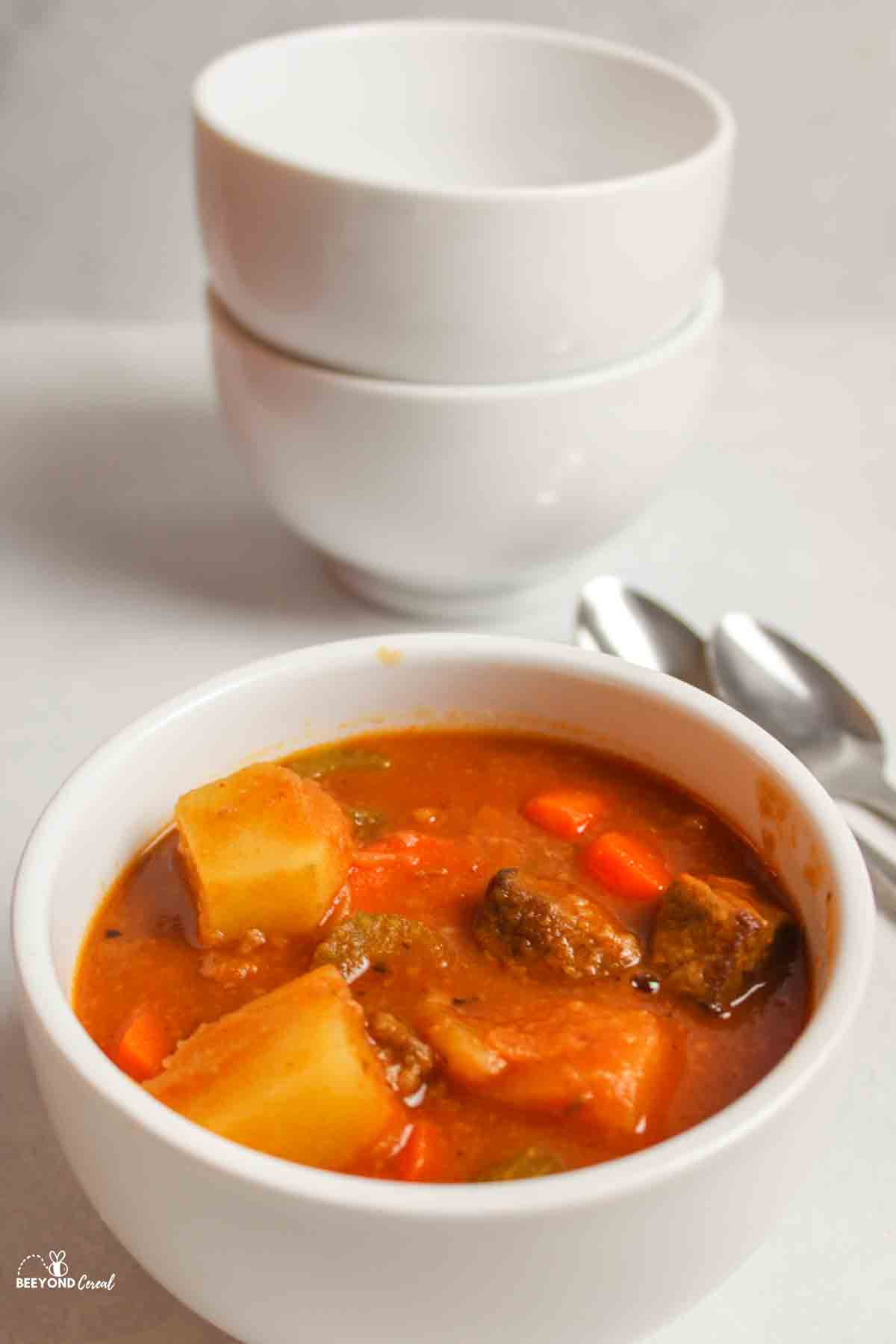a white bowl filled with beef stew with spoons and empty stacked bowls in background