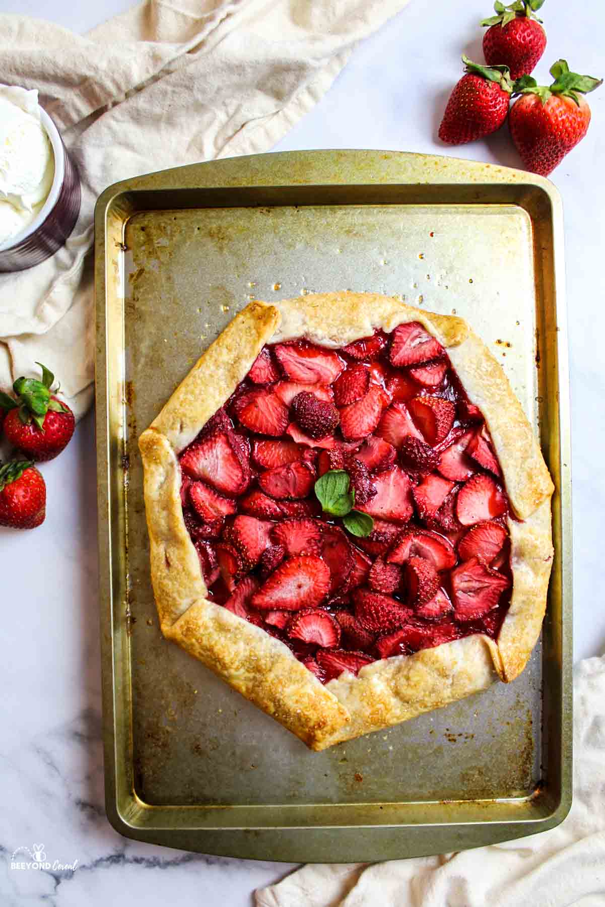 aerial view of strawberry galette on a baking sheet with fresh berries, cream, and kitchen towel around it