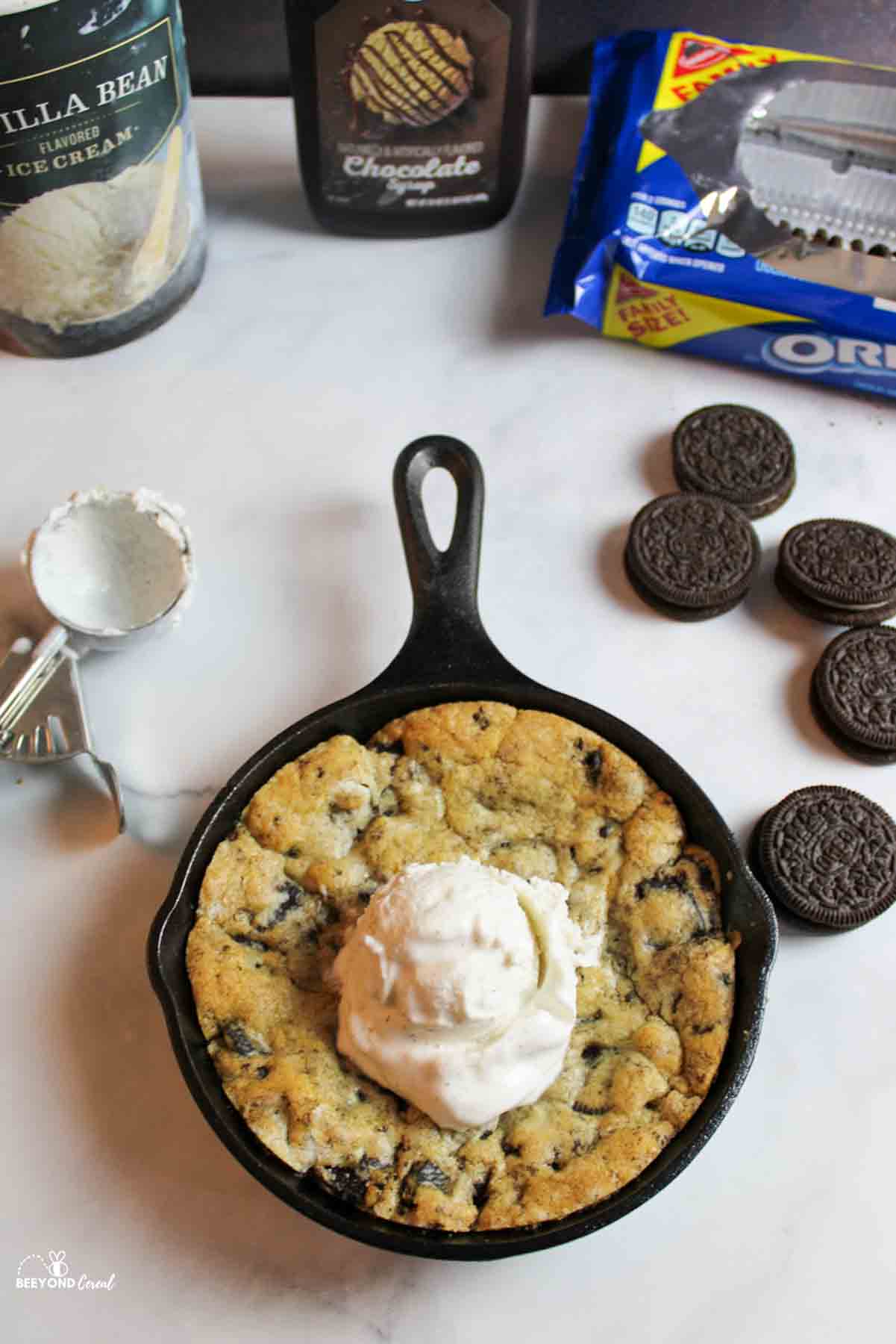 an oreo pizookie in a cast iron skillet with ice cream scoop on top. Use scooper to the side and more food items such as ice cream container, chocolate syrup bottole and oreos in and out of package around the skillet cookie