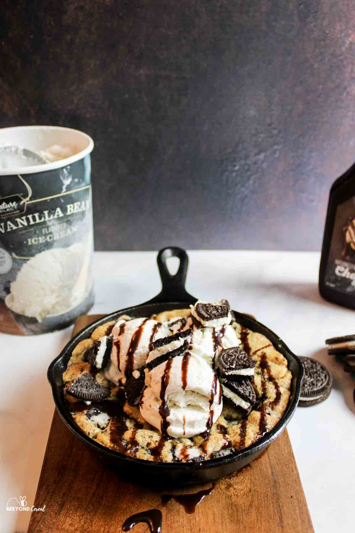 a chocolate syrup and oreo topped vanilla ice cream scoop above a bakedoreo pizookie with ice cream and chocolate syrup bottle in background
