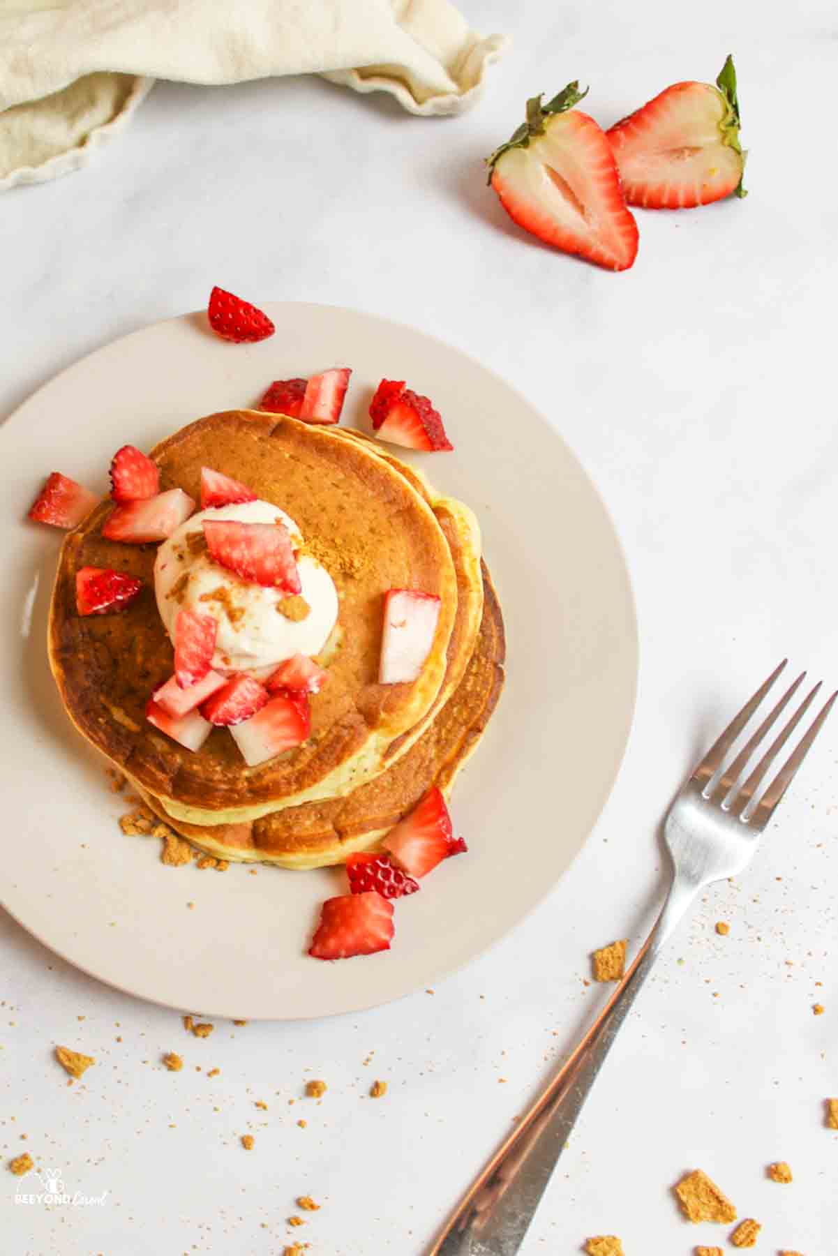 Plate of strawberry cheesecake pancakes with fork, sliced strawberry aside.