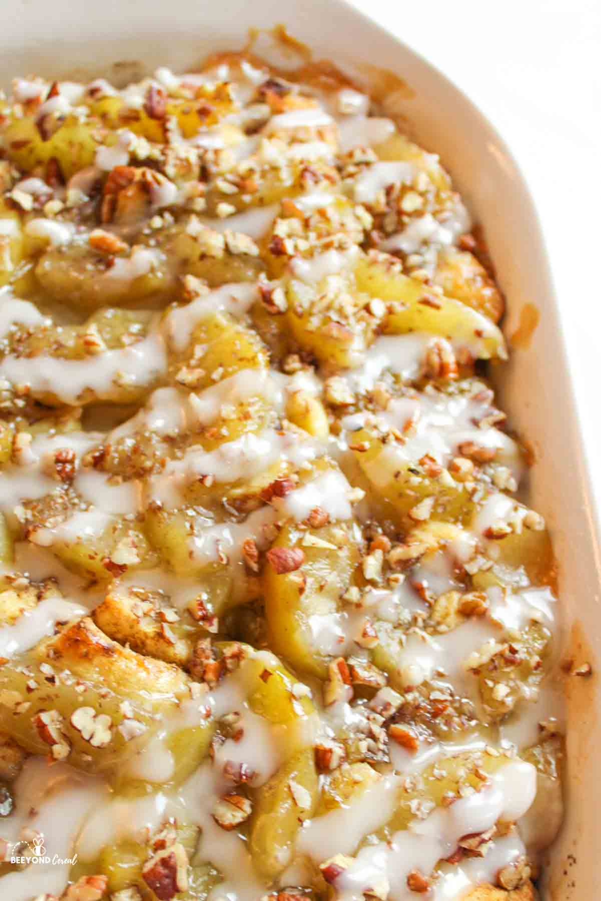 an upclose view of half the pan of cinnamon roll apple cobbler with icing and pecans on top