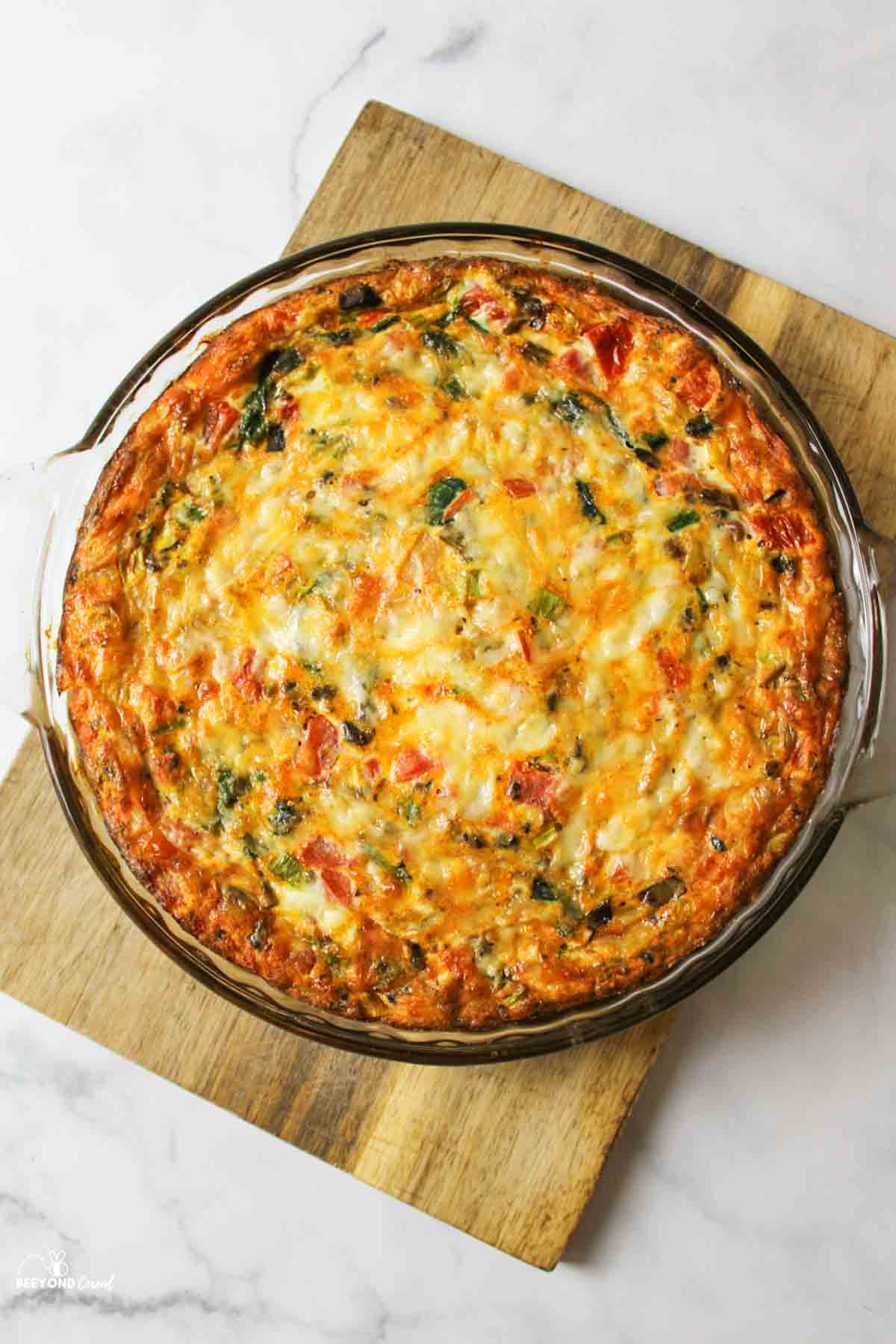 baked veggie quiche in a glass pie plate on a wooden cutting board.