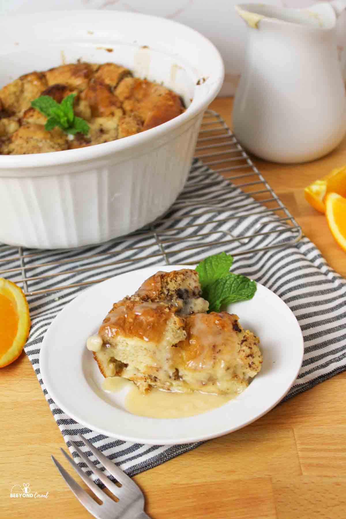 a slice of bread pudding on a white plate with more in a round casserole dish behind it