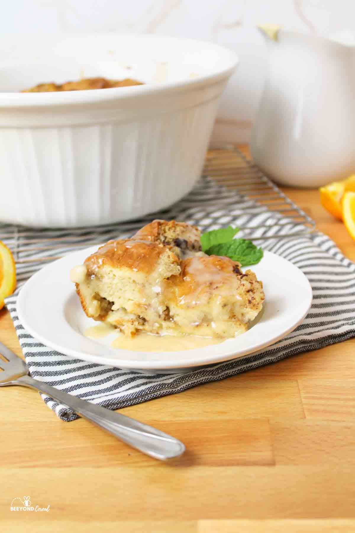 Slice of bread pudding on white plate, casserole dish behind, fork.