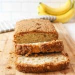 sliced banana bread on a cutting board with ripe bananas in background.