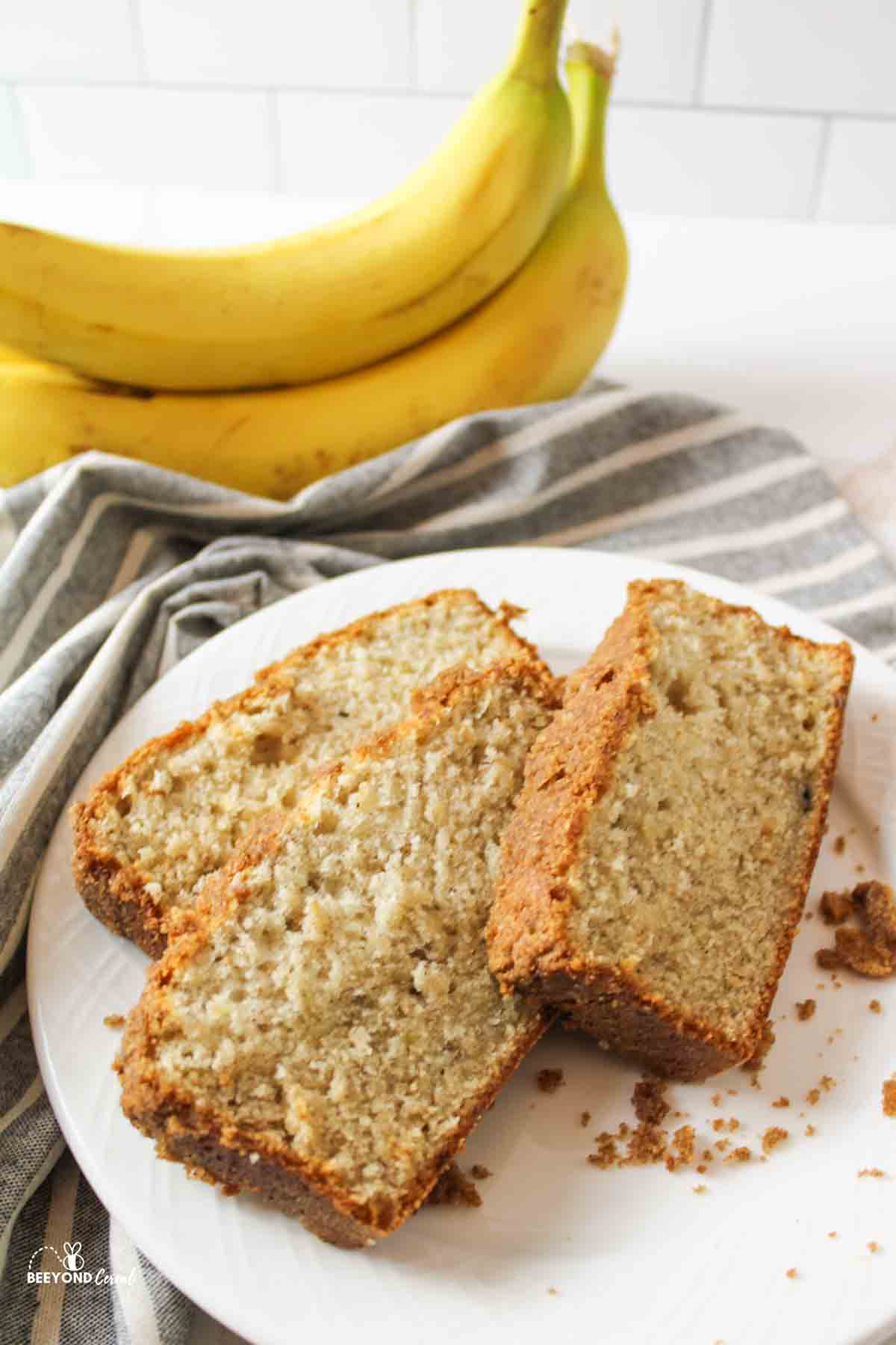 three slices of cinamon crumble banana bread on a plate next to a striped towel and fresh bananas