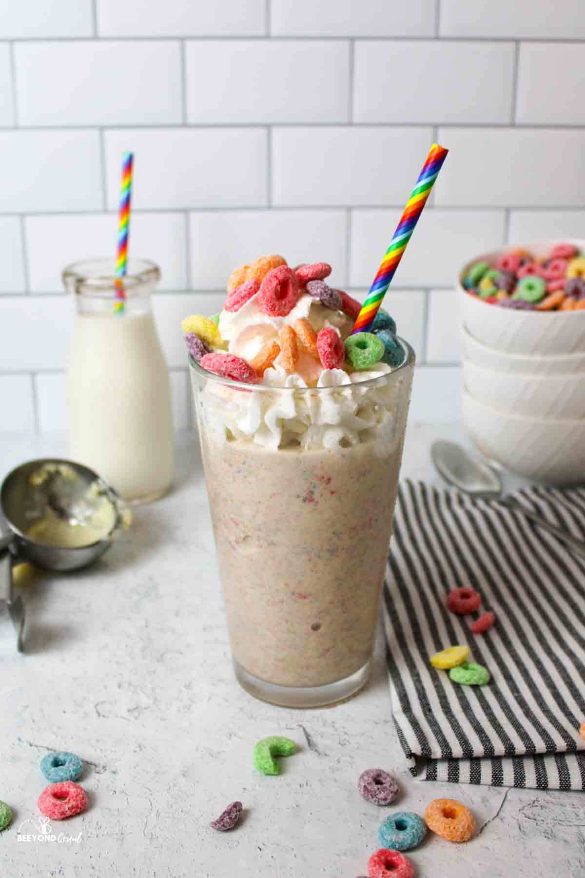 a glass of milkshake with cereal and scattered fruit loops around it.