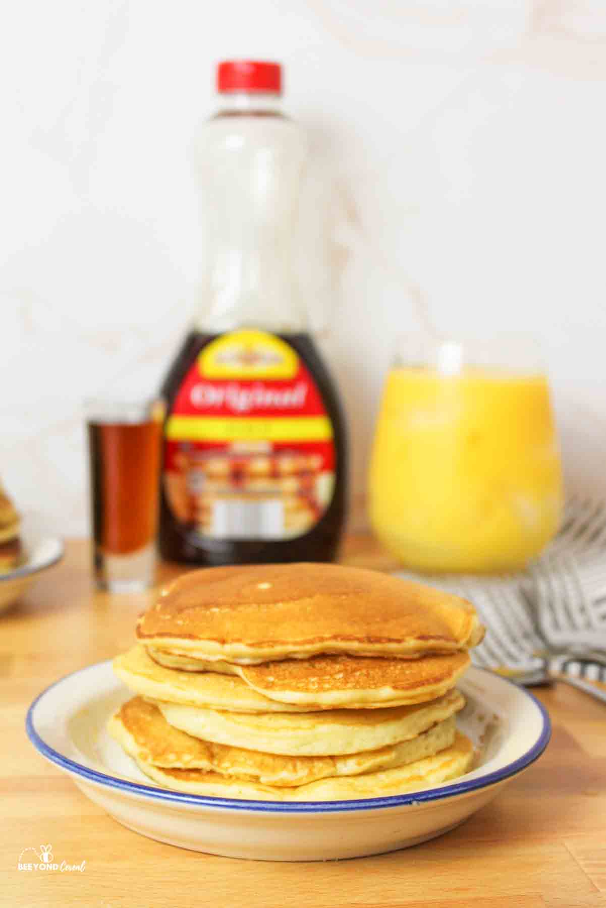 a stack of golden pancakes on a blue rimmed plate with syrup and orange juice in background.