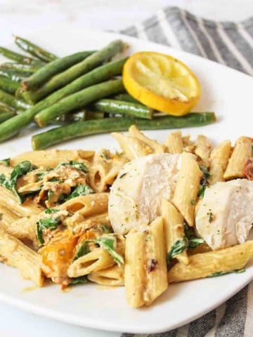 an upclose view of roasted garlic and tomato pasta with chicken and a pile of green beans and lemon slice in back