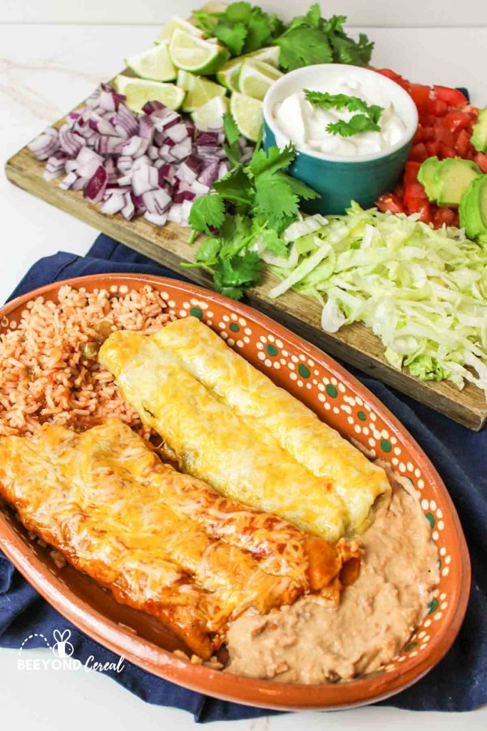 an oval plate filled with enchiladas and sides with toppings arranged on a board in background
