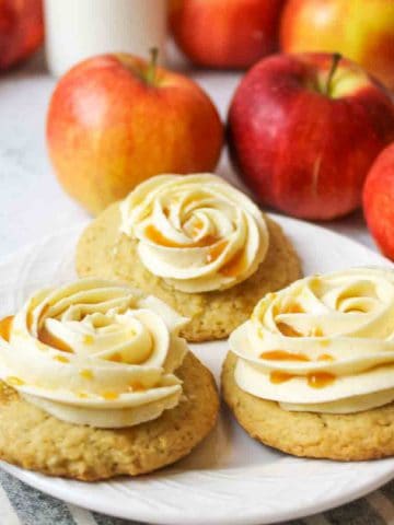 three caramel buttercream topped apple cider cookies next to fresh red apples on a white plate.