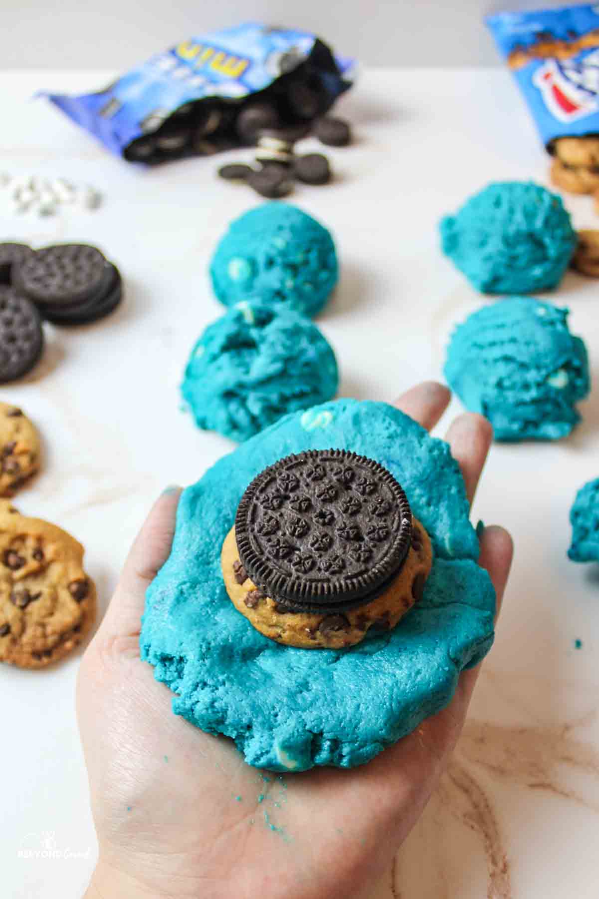 a hand holding a large blue dough topped with a chips ahoy cookie and oreo.