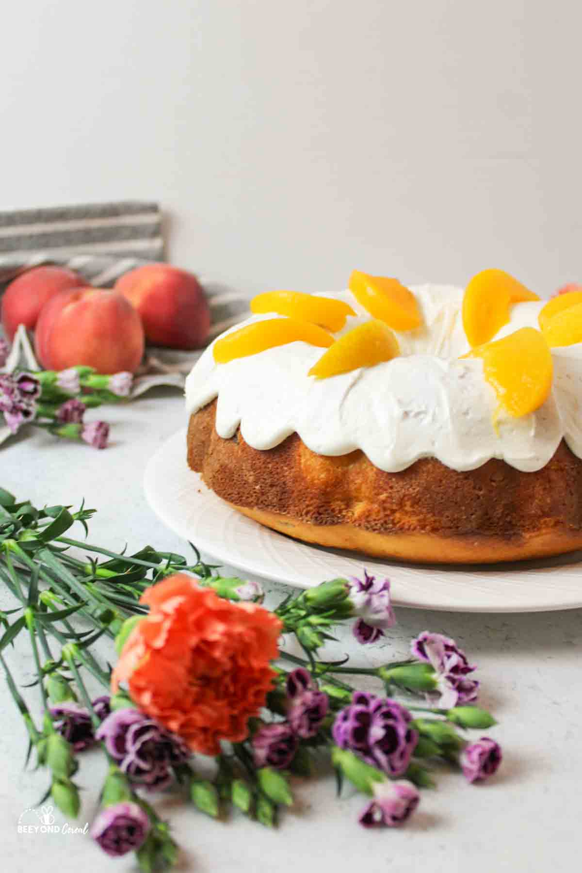 a side view of a peaches and cream bundt cake with fresh flowers and peaches to the side