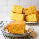 a slice of cornbread on a blue rimmed plate with more stacked pieces of cornbread in the background