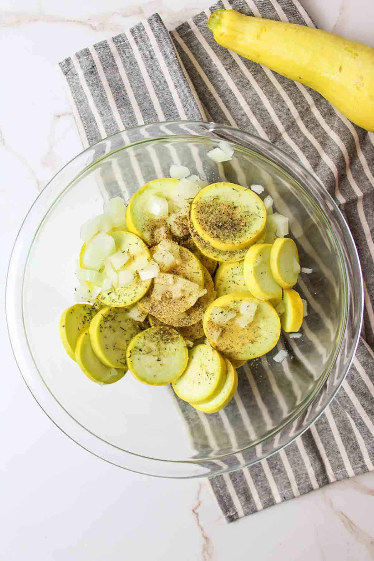 sliced yellow squash and diced onions in a bowl with seasoning on top.