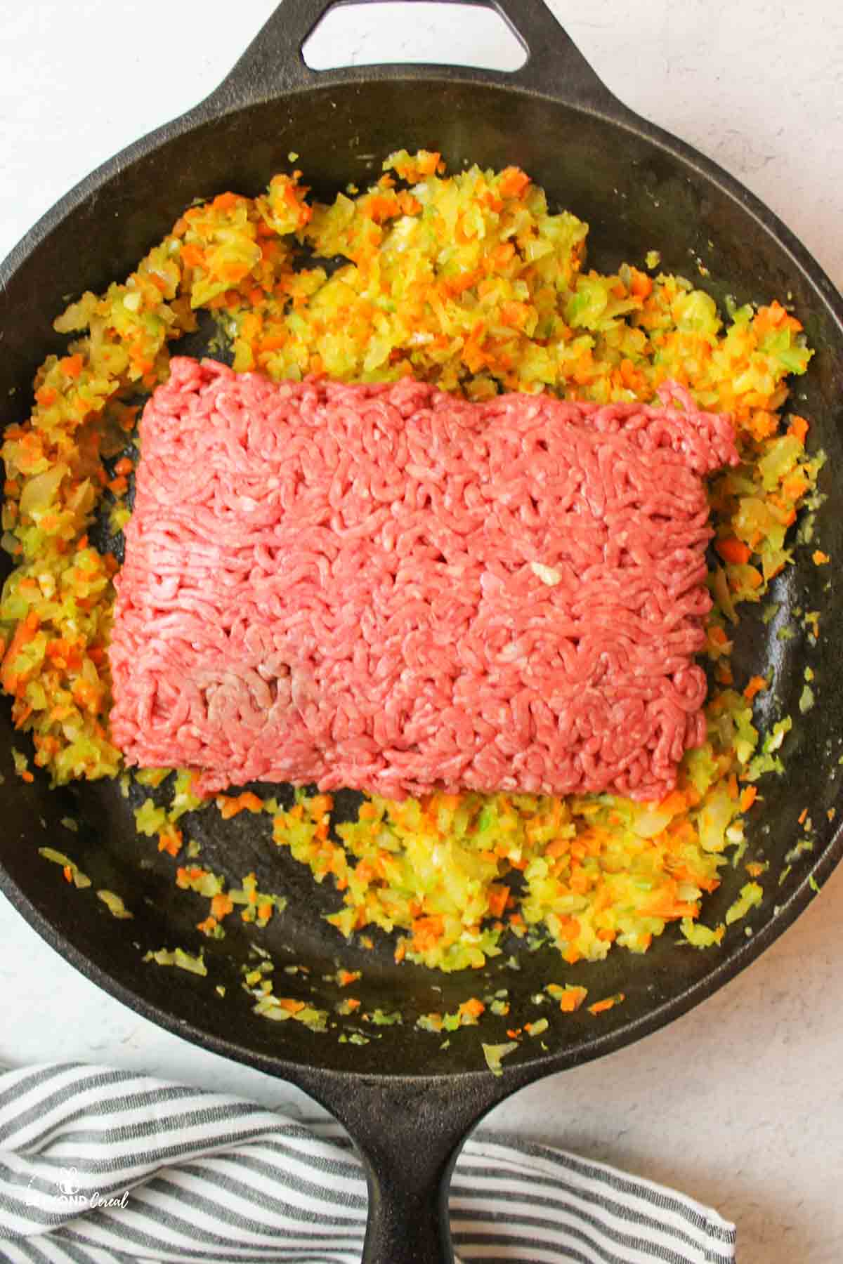 raw ground beef added to a skillet of cooked carrots, onion, garlic, and celery