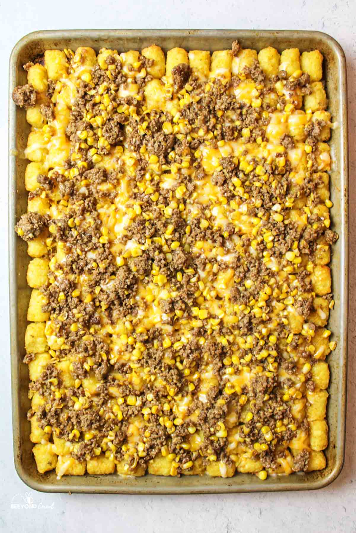 cheese and meat with corn on top of tater tots on baking sheet