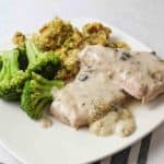 two crockpot cooked porkchops smothered in mushroom gravy and served next to broccoli and stuffing on a white dinner plate.