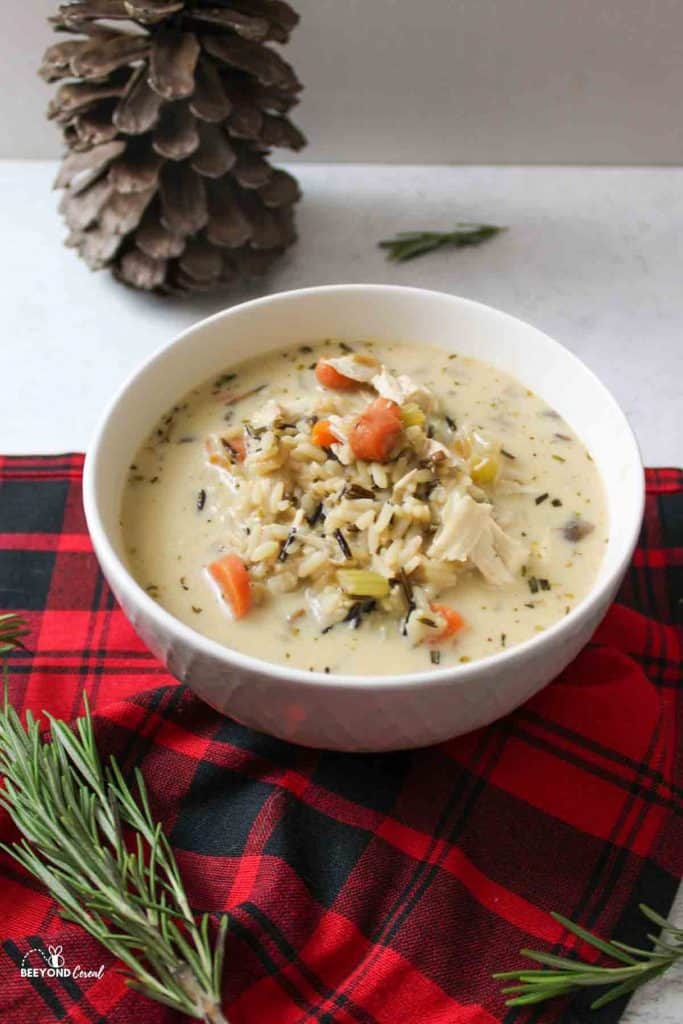a bowl of turkey wild rice soup next to fresh rosemary, a red plaid towl and a pinecone in background