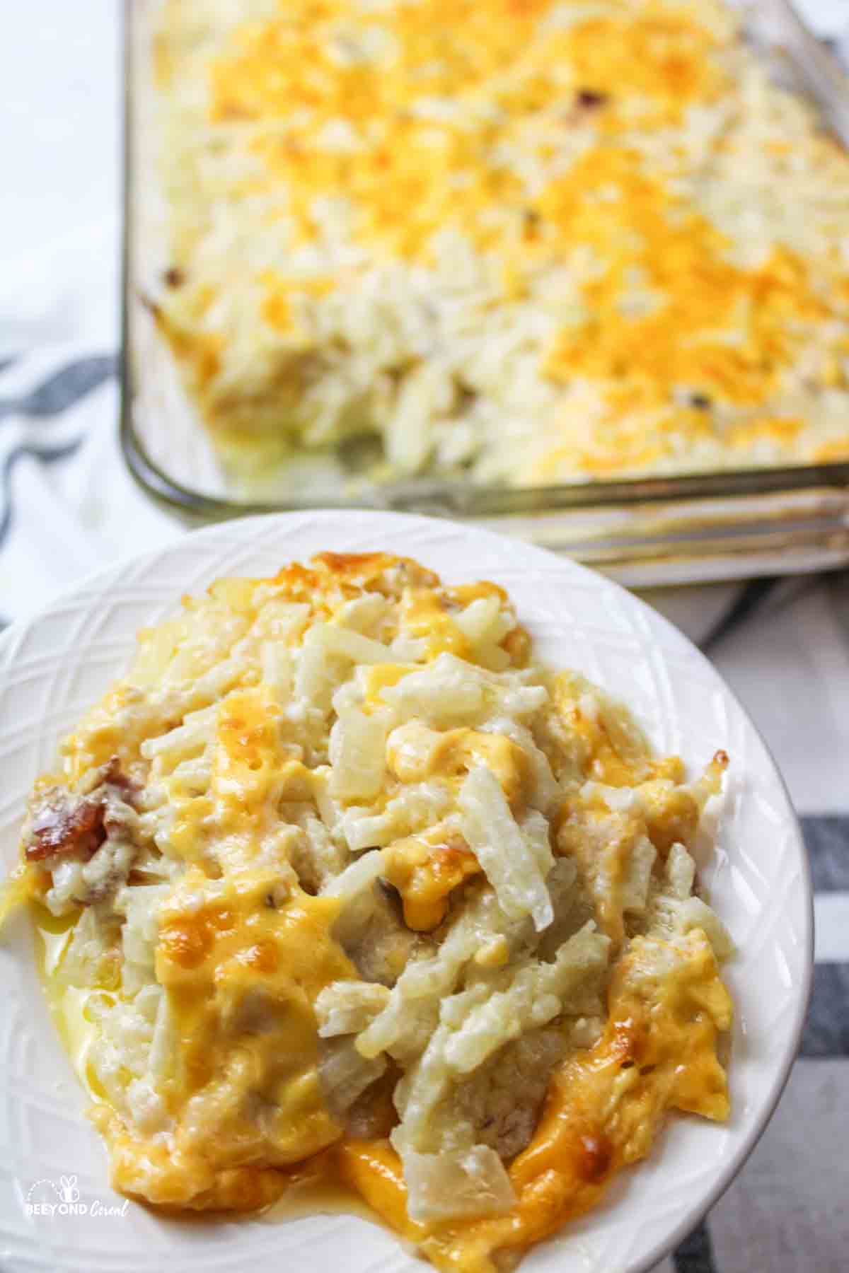 a serving of cheesy hashbrown casserole on a plate with more in a baking dish in background