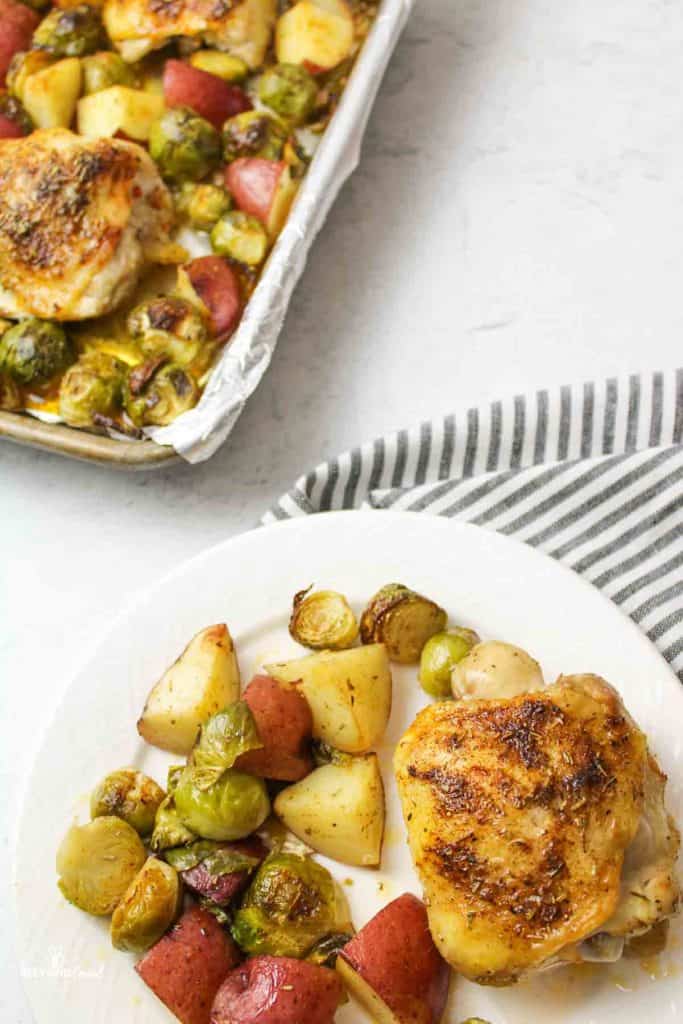 a dinner plate filled with brussel sprouts, red potatoes and cooked chicken thighs with more in a sheet pan to the side