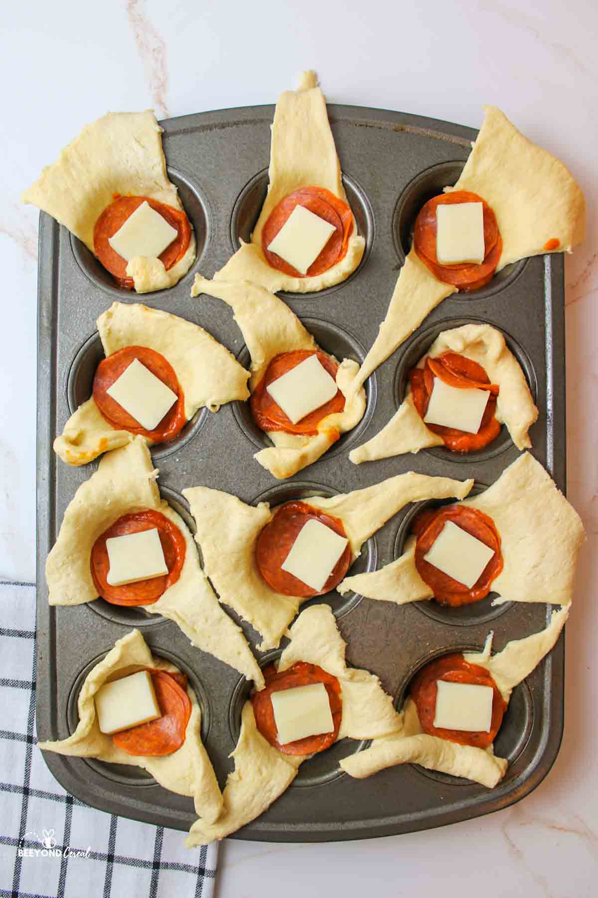 crescent rolls in muffin cups with a dollop of pizza sauce in each center and then topped with pepperoni slices and a chunk of mozzarella cheese