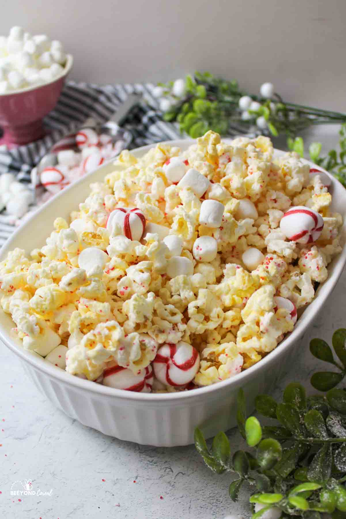 a side view of a white oval dish filled with white chocolate peppermint popcorn, mini marshmallows, and peppermint balls