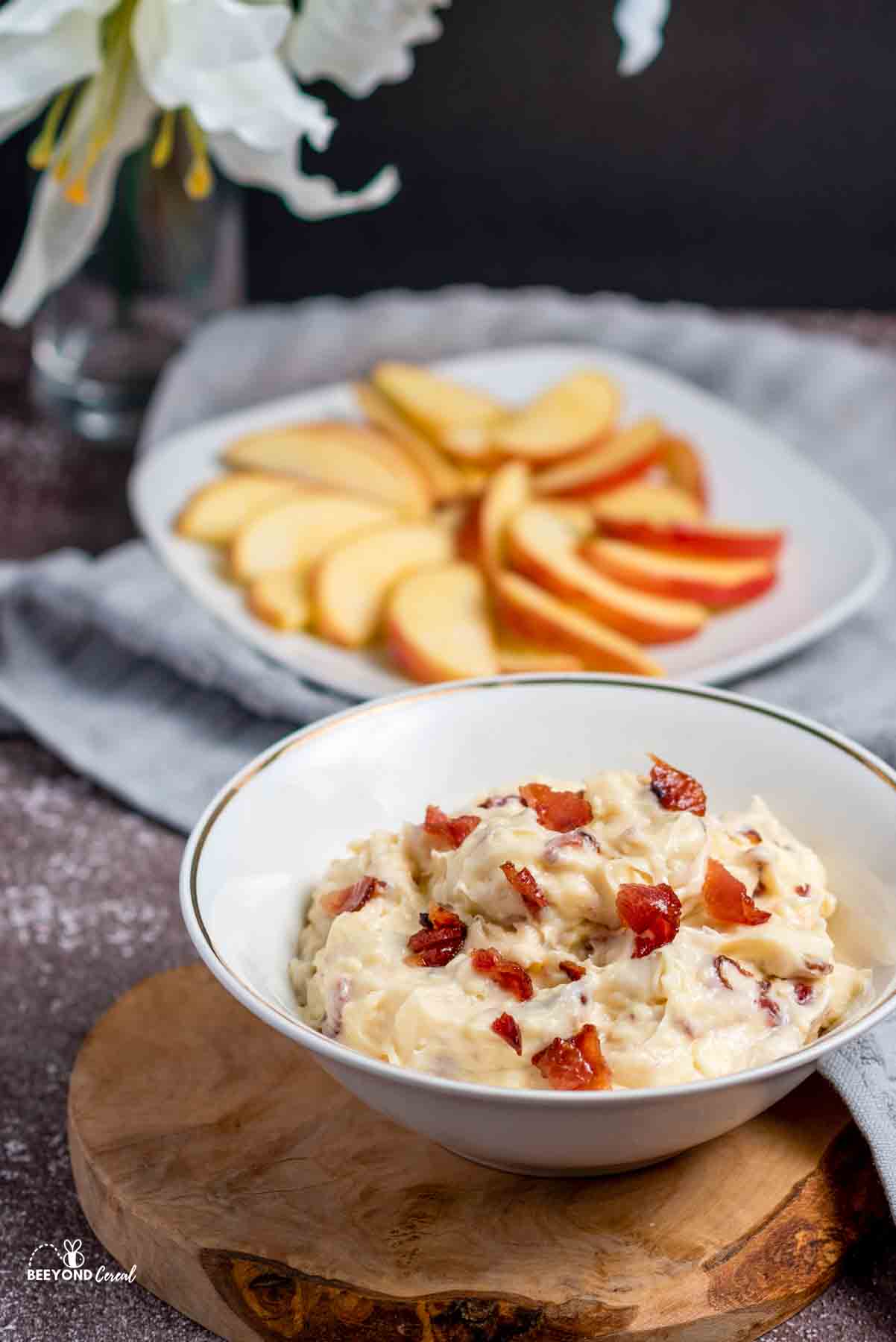a bowl of maple bacon dip topped with crumbled bacon and a plate of apple slices in the background