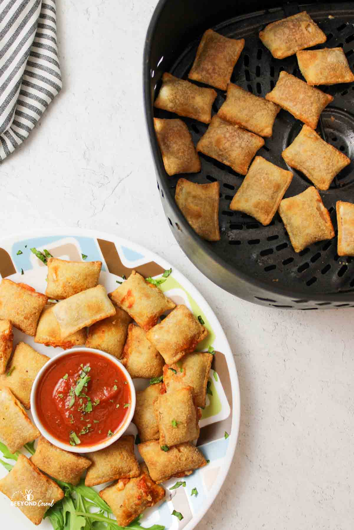 a plate of pizza rolls with marinara sauce next t an air fryer basket with more pizza rolls inside