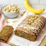 a sliced loaf of pistachio banana bread with a bowl of pistachios and a banana in background