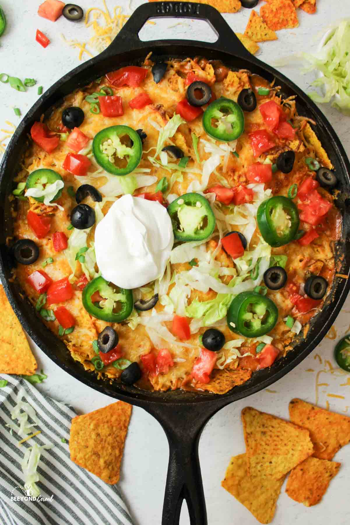walking taco casserole in a cast iron and garnished with jalapenos, lettuce, tomatoes, olives and sour cream