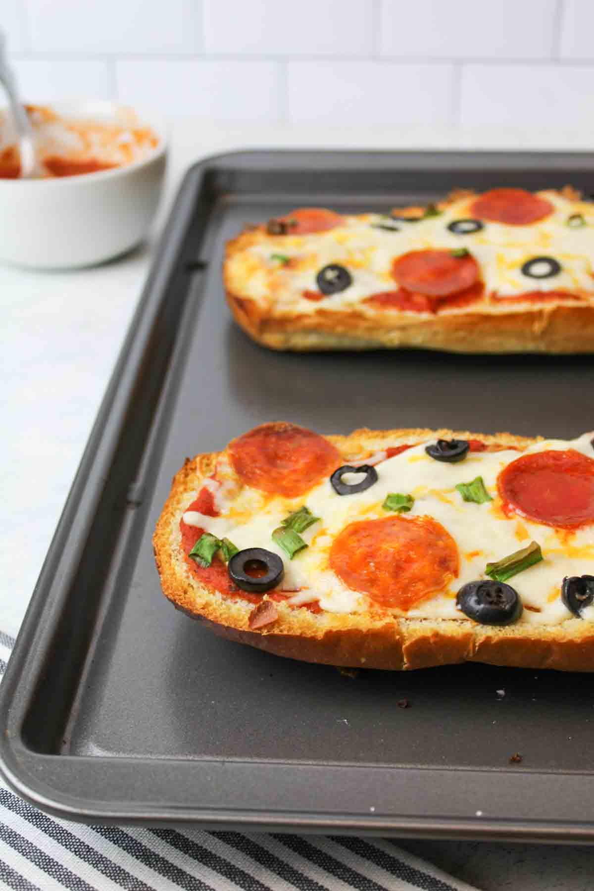 two baked halves of french bread pizza on a baking sheet with a bowl of sauce in the background