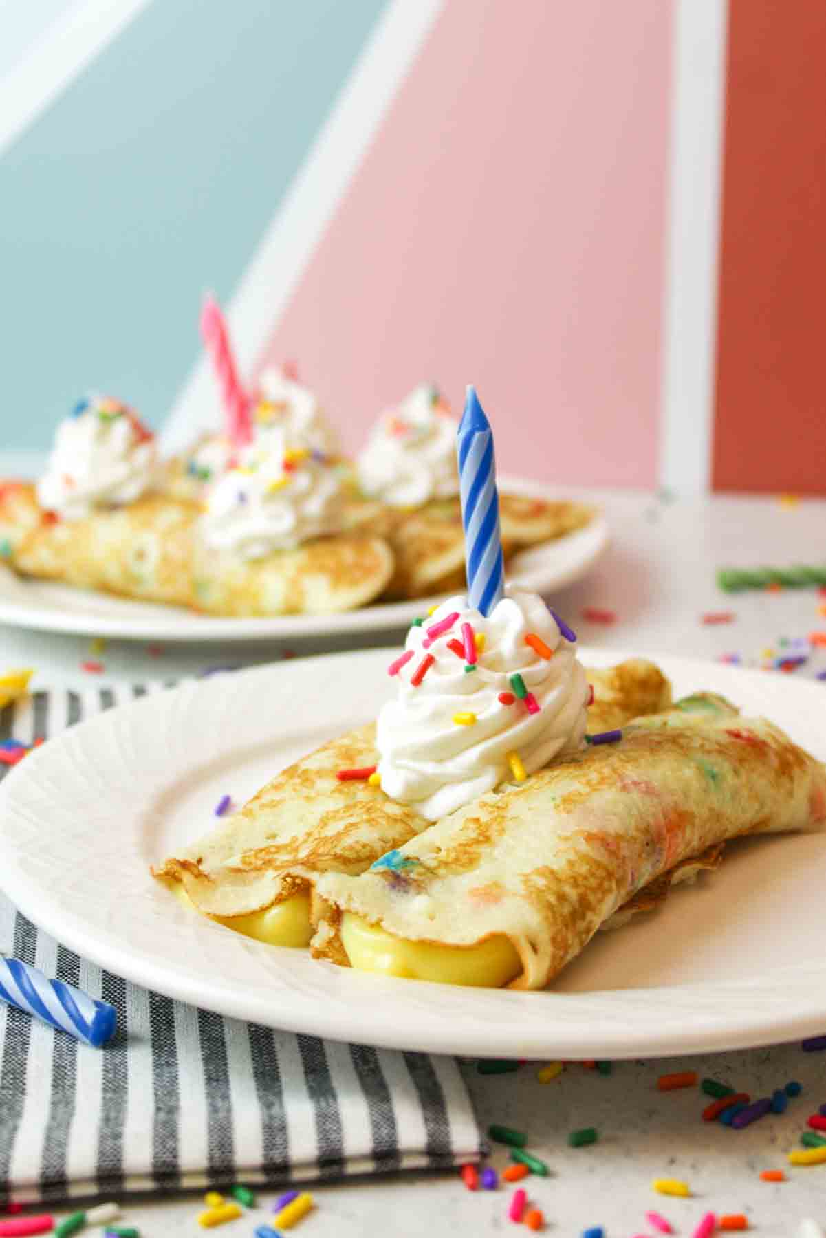 birthday cake crepes with whipped cream, sprinkles, and candles served on white plates.