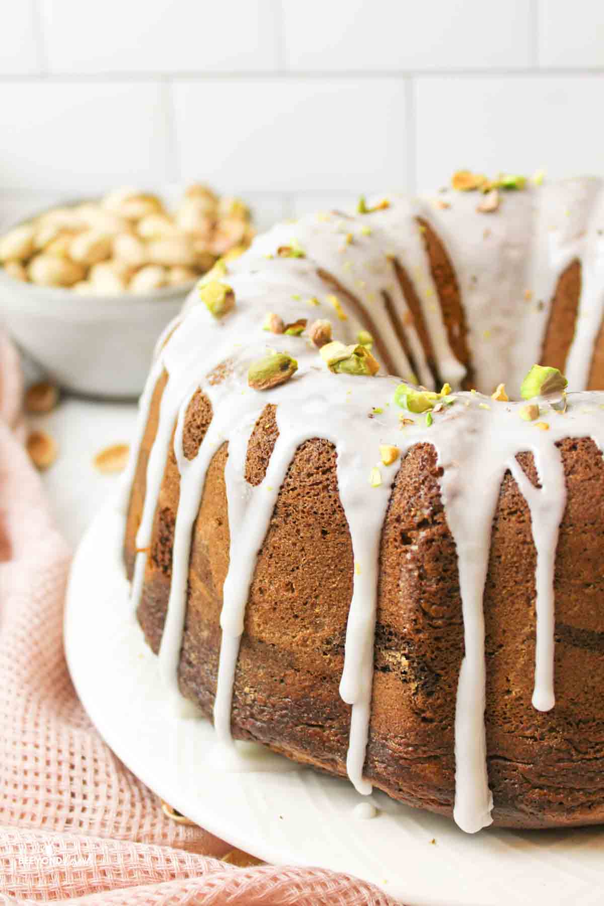 a side view of a glazed Chocolate Pistachio Bundt Cake with bowl of pistachios in backround