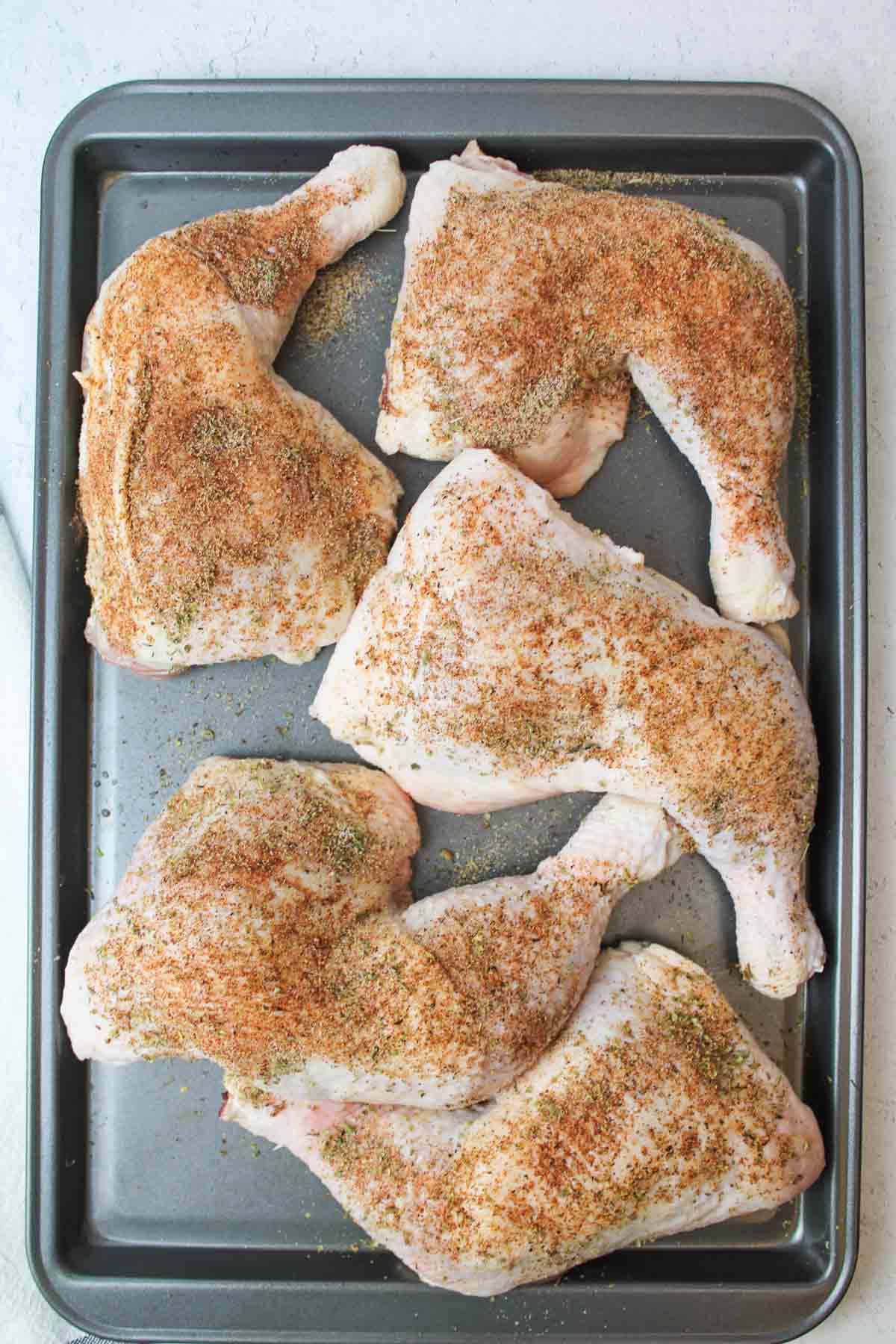 chicken quarters on a baking sheet with seasonings sprinkled on top.