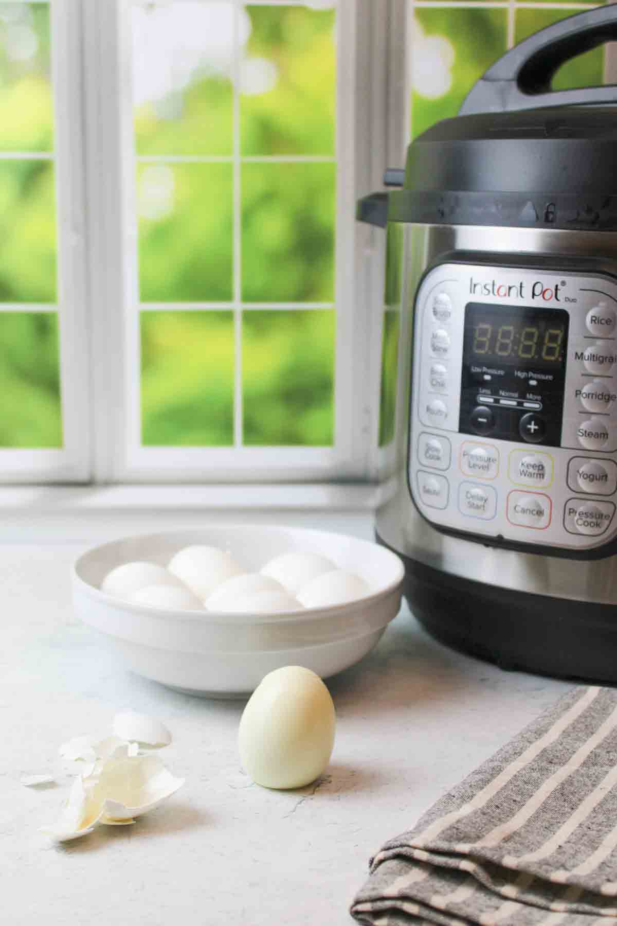 Bowl of eggs next to Instant Pot, one peeled and standing in front.