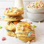 a stack of lucky charms cookies with one cookie resting beside it and a bowl of cereal in the background