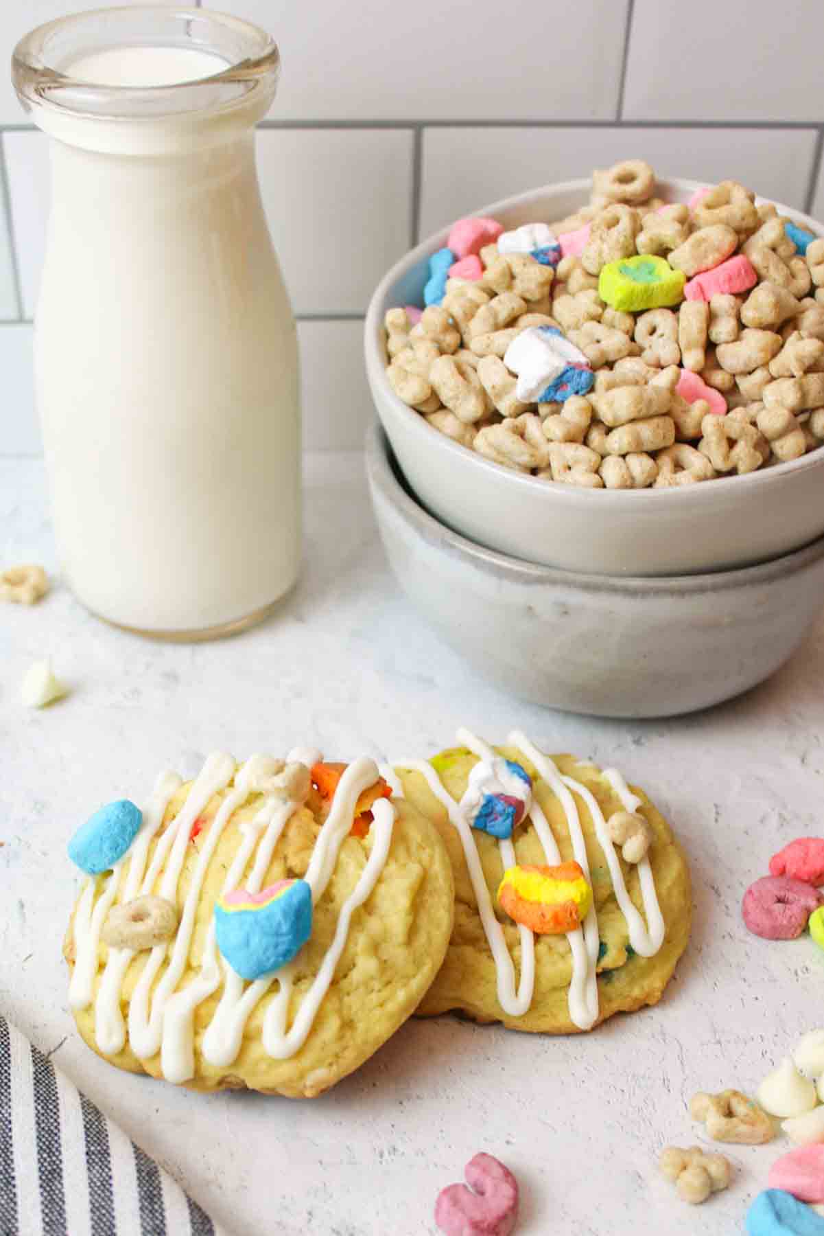 two lucky charms cookies next to a glass of milk and bowl of cereal.