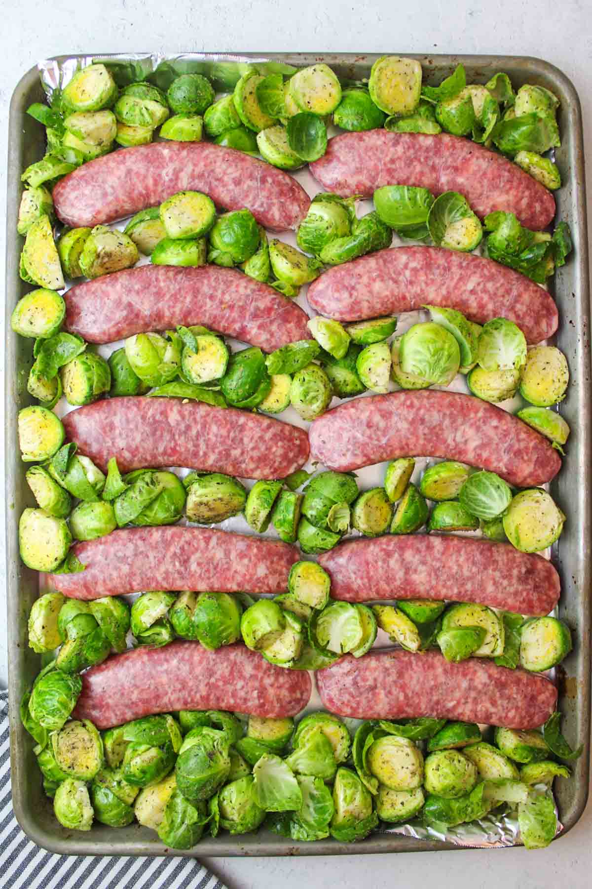 raw sausages on a baking sheet with halved brussel sprouts around them.