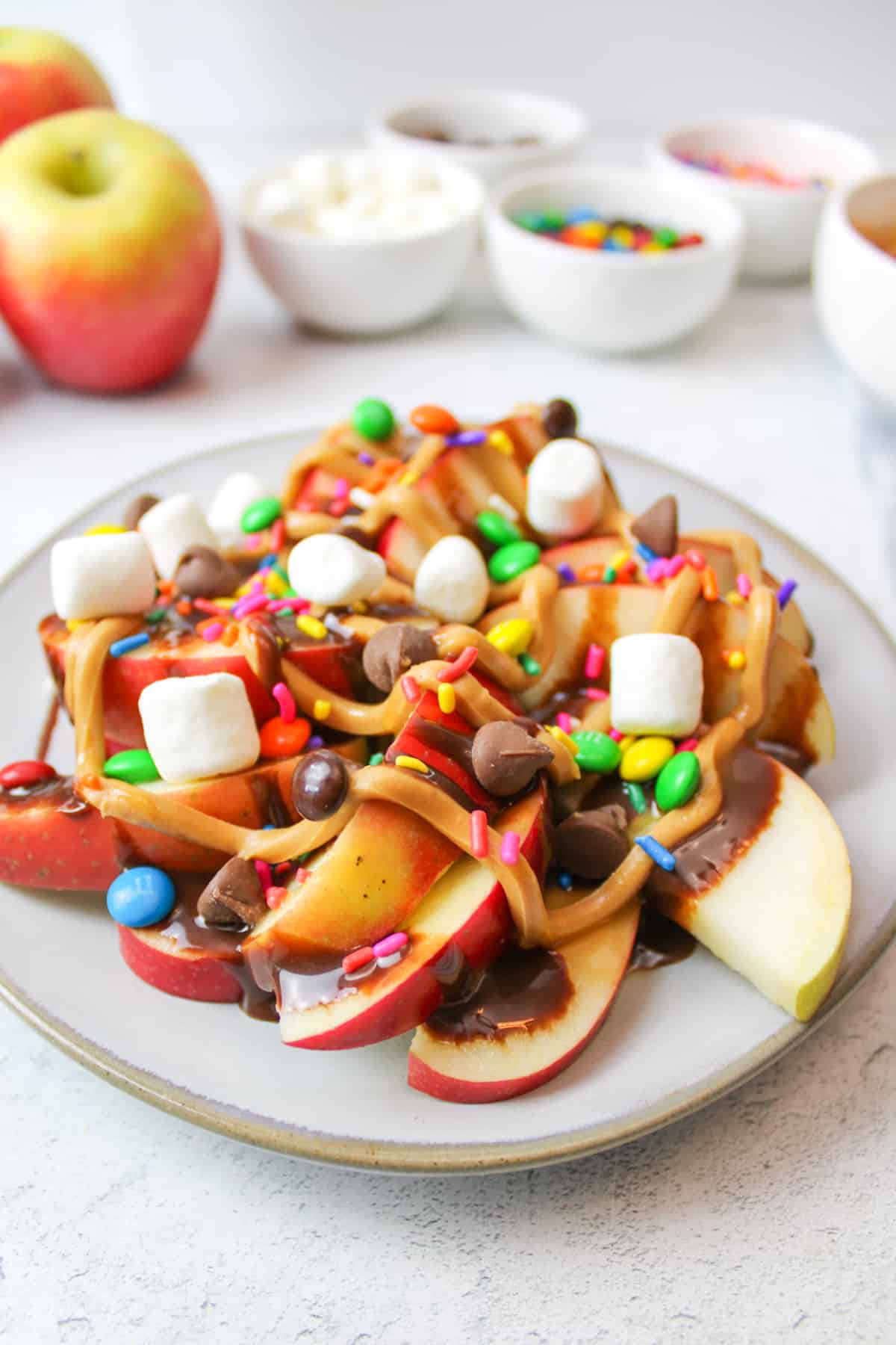 side view of apple nachos on a plate with bowls filled with toppings in background