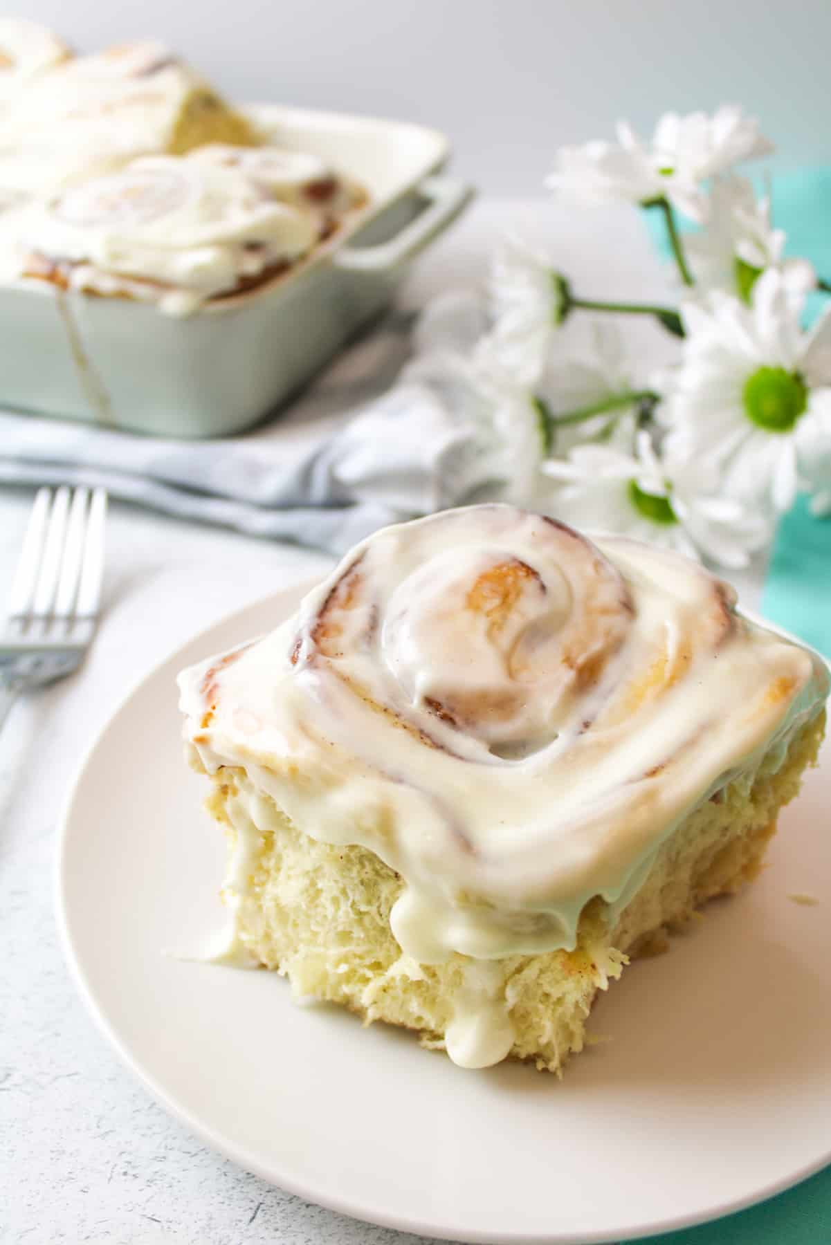 a frosted cinnamon roll on a plate with more rolls in background near fresh flowers and a fork