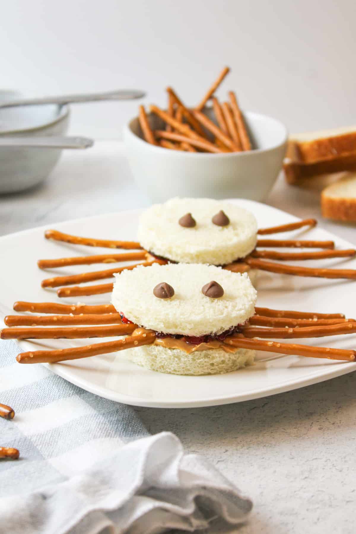 two spider sandwiches on a plate with a bowl of pretzel sticks in background.