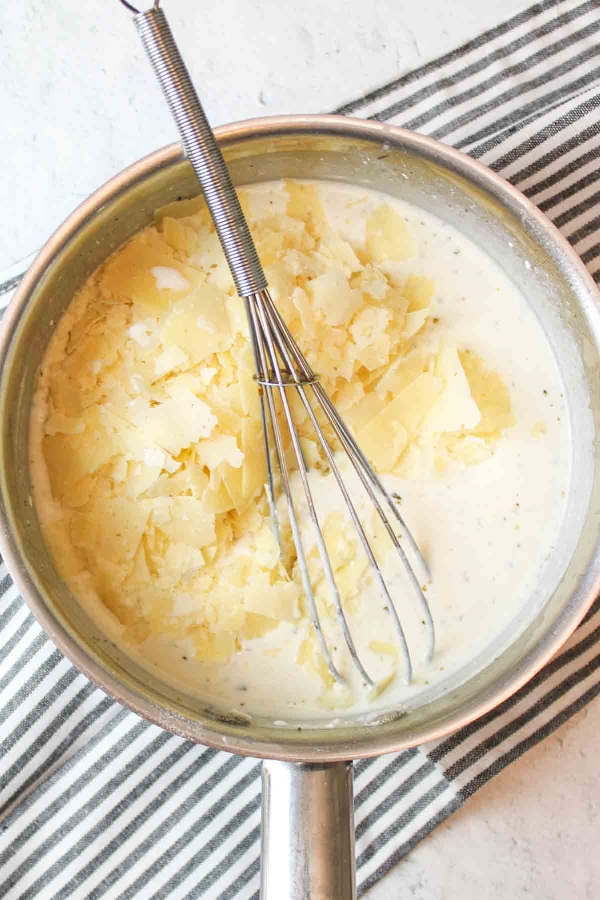 a whisk in a saucepan of cream cheese alfredo and fresh parmesan added to it.