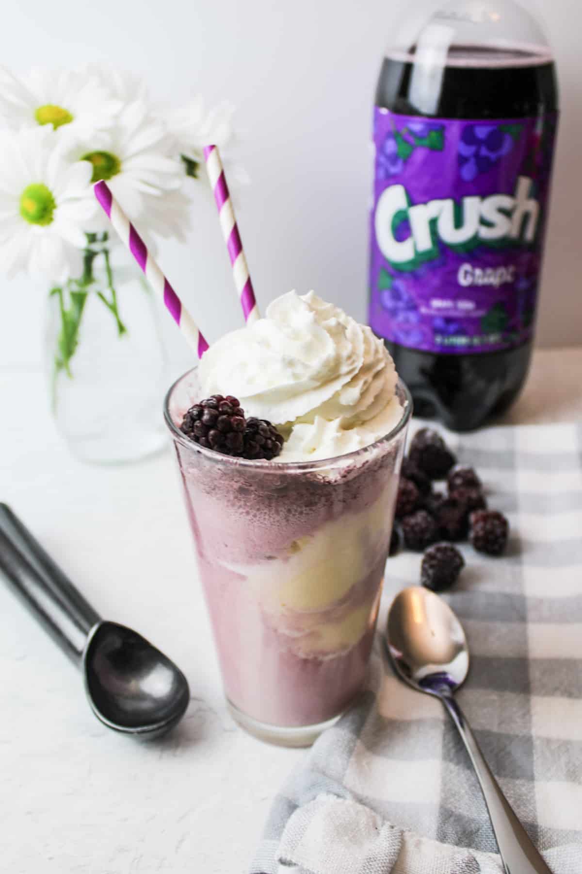 a glass cup full of purple cow float with spoons and berries to the side.