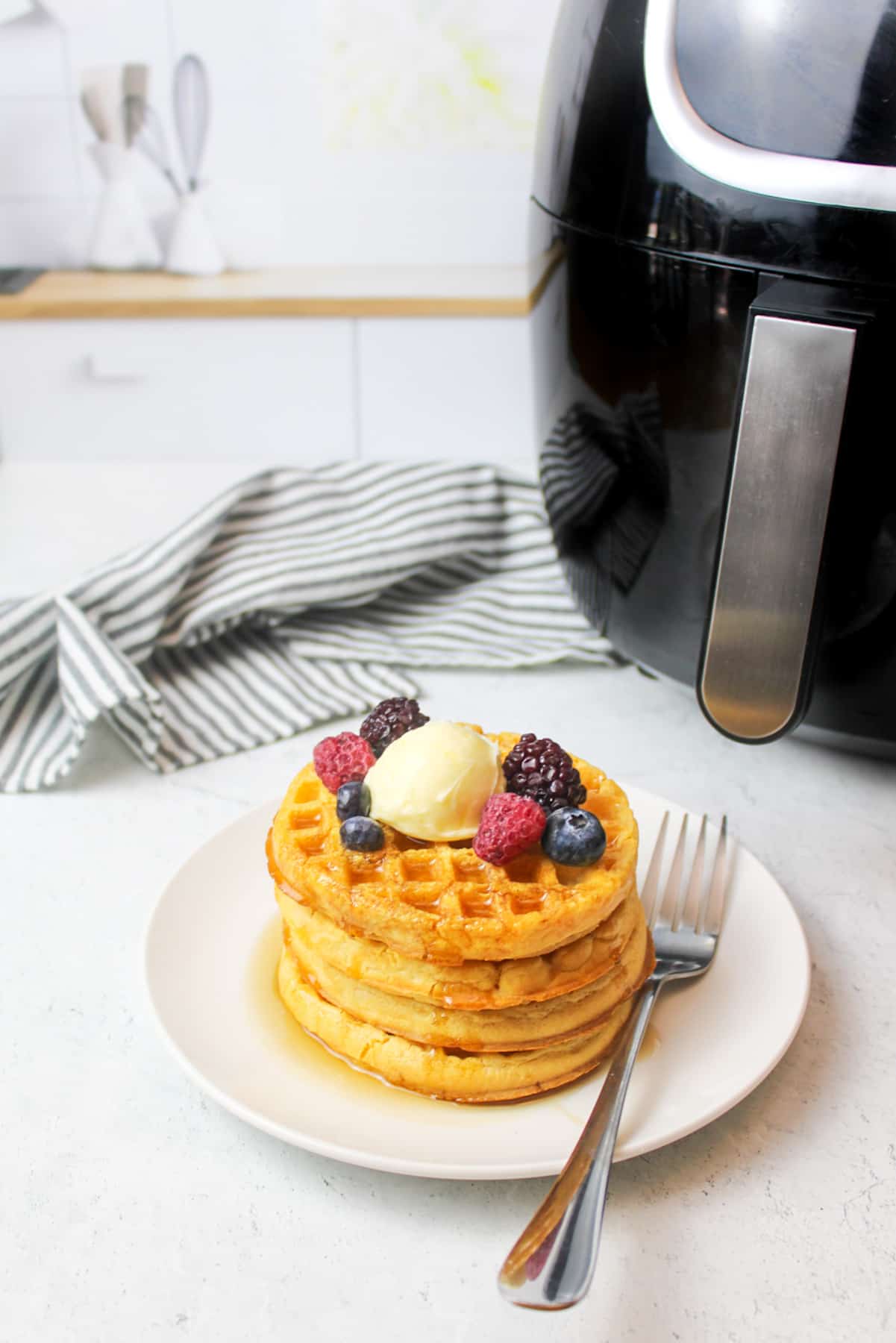 air fryer next to cooked frozen waffles stacked on a plate with berries and a fork.